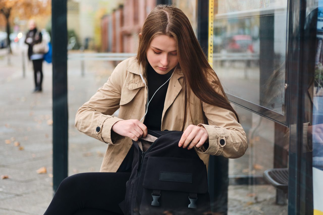 Studentka ze słuchawkami czeka na przystanku i szuka czegoś w plecaku